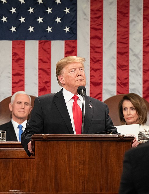 President Trump Stands At A Podium In Front Of A Large American Flag. Behind Him Sit Vice President Pence And Speaker Of The House Pelosi.