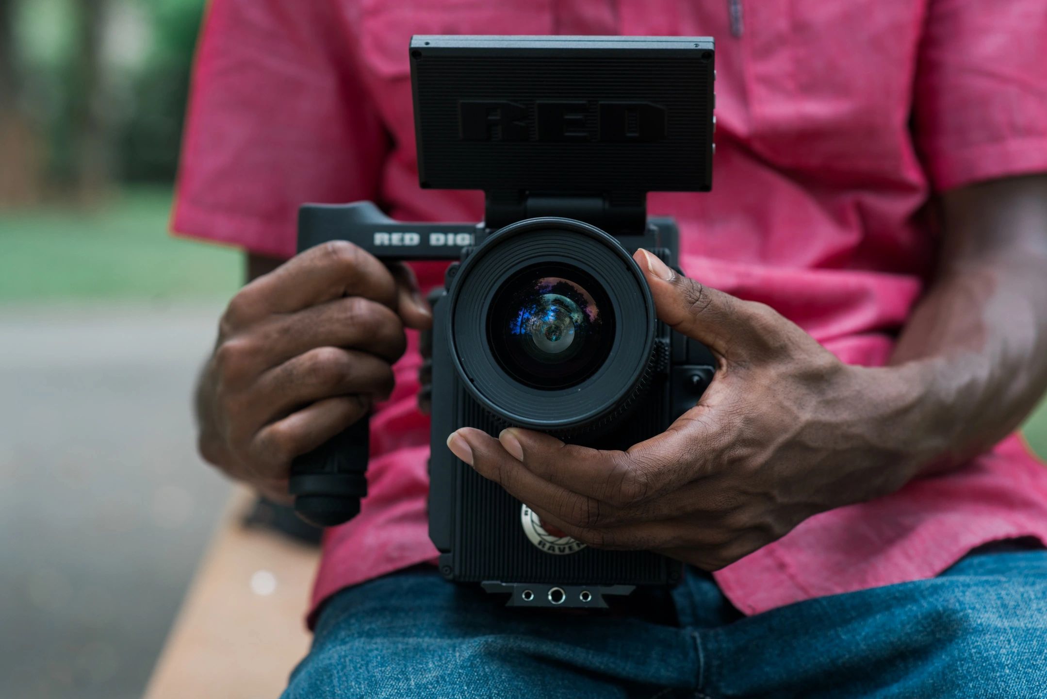 Close Up Image Of A Camera Being Held By What Appears To Be An African American Man Wearing A Red Shirt. Only The Camera Is Visible, You Do Not See The Person's Head Or Legs.