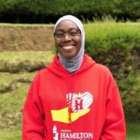 Image Of An African American Woman Wearing A Red Sweatshirt, Glasses, And A Head Scarf. She Is Standing Outside And Smiling At The Camera.