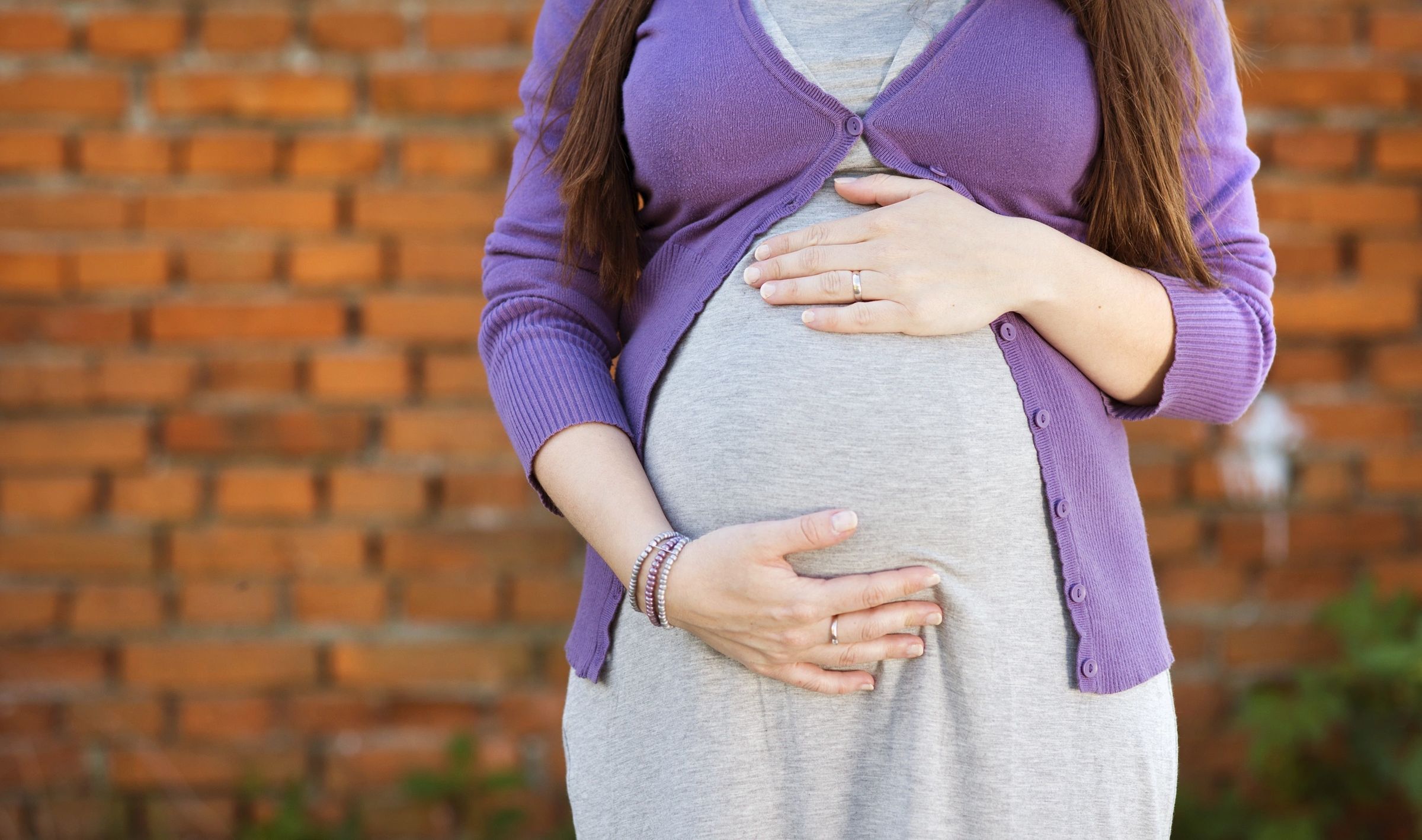 Image Of The Midsection Of A Pregnant Woman Wearing A Grey Dress And Purple Sweater. Her Hands Are On Her Belly. Neither Her Head Nor Legs Are Showing.