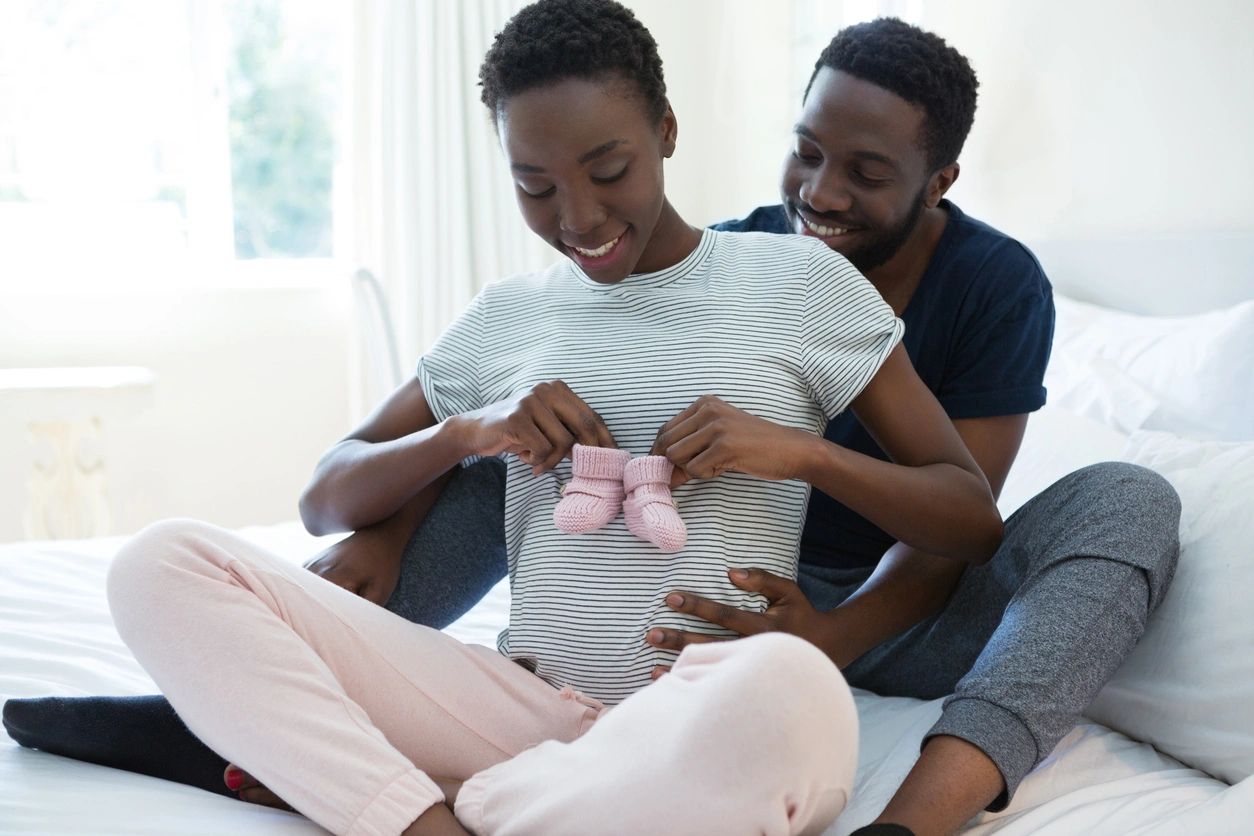 Image Of An African American Couple Sitting On The Floor. The Woman Is Pregnant And Holding Up Two Baby Booties And The Man Is Sitting Behind Her Smiling.