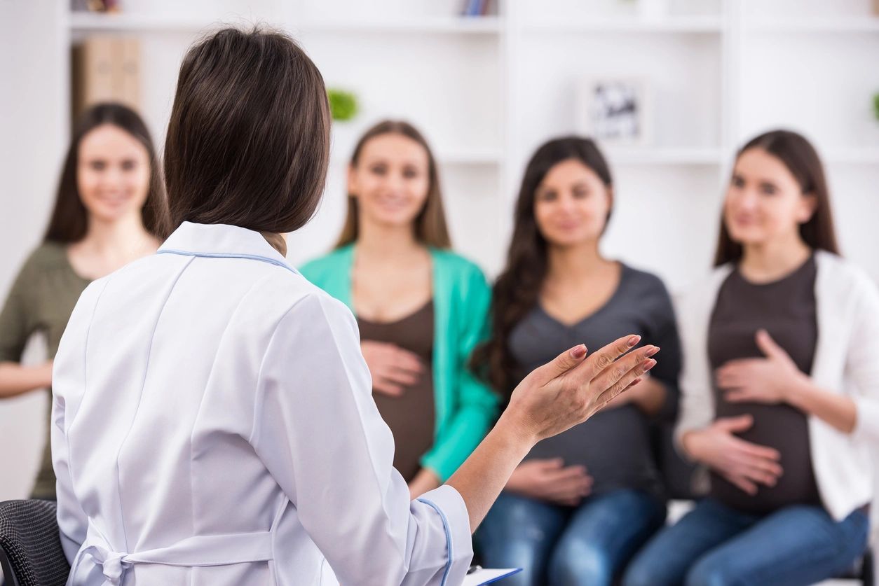 Image Of A Doctor (seen From The Back, Wearing A White Coat) Speaking To Four Women, All Of Whom Appear To Be Pregnant