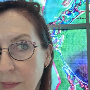 A Pale-skinned Woman With Dark-red Hair Looks Skeptically Sideways At A Stained Glass Microbe.
