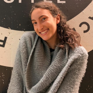 Image Of A White Woman Wearing A Grey Sweater. She Is Seen From Her Waist Up And Is Smiling.