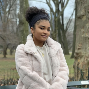 Picture Of A Young Black Woman With Her Hair Up In A Bun. She Is Wearing A Fuzzy White Jacket.