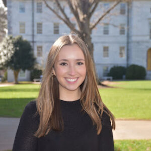 A Young Caucasian Woman With Long Blonde Hair Stands Against A Campus Backdrop Of Green Grass And White Stone Buildings.