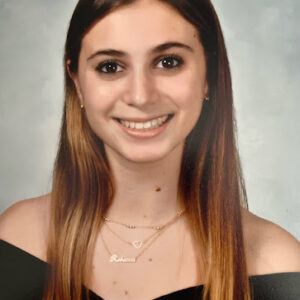 A Woman With Medium Skin Tone And Dark Eyes Is Pictures Against A Gray Photographer's Background. She Wears A Black Drape Esposing Her Shoulders And Several Narrow Gold Necklaces. Her Long Hair Is Dark At Her Parting And Amber Toward The Ends.