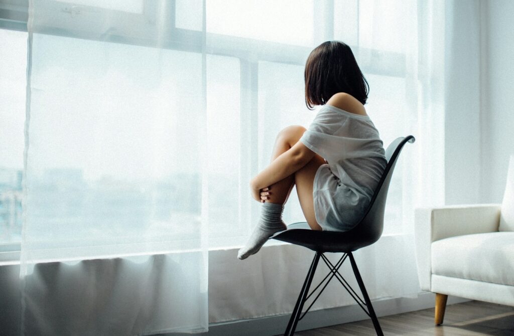 Seen from behind, a dark-haired woman in a white gown curls up in a chair and stares through a curtained window.