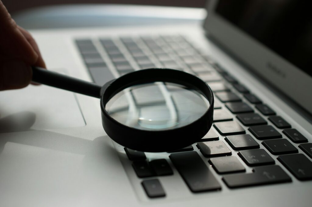 A hand holds a magnifying glass over a laptop keyboard.