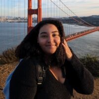 A Young Black Woman Wearing A Long-sleeved Dark Top Smiles At The Viewer. She Is Standing On A Bluff Above San Francisco's Golden Gate Bridge.