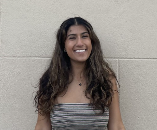 A young woman with center-parted brown hair smiles at the camera against a stone-block wall. She is wearing a striped tube top.