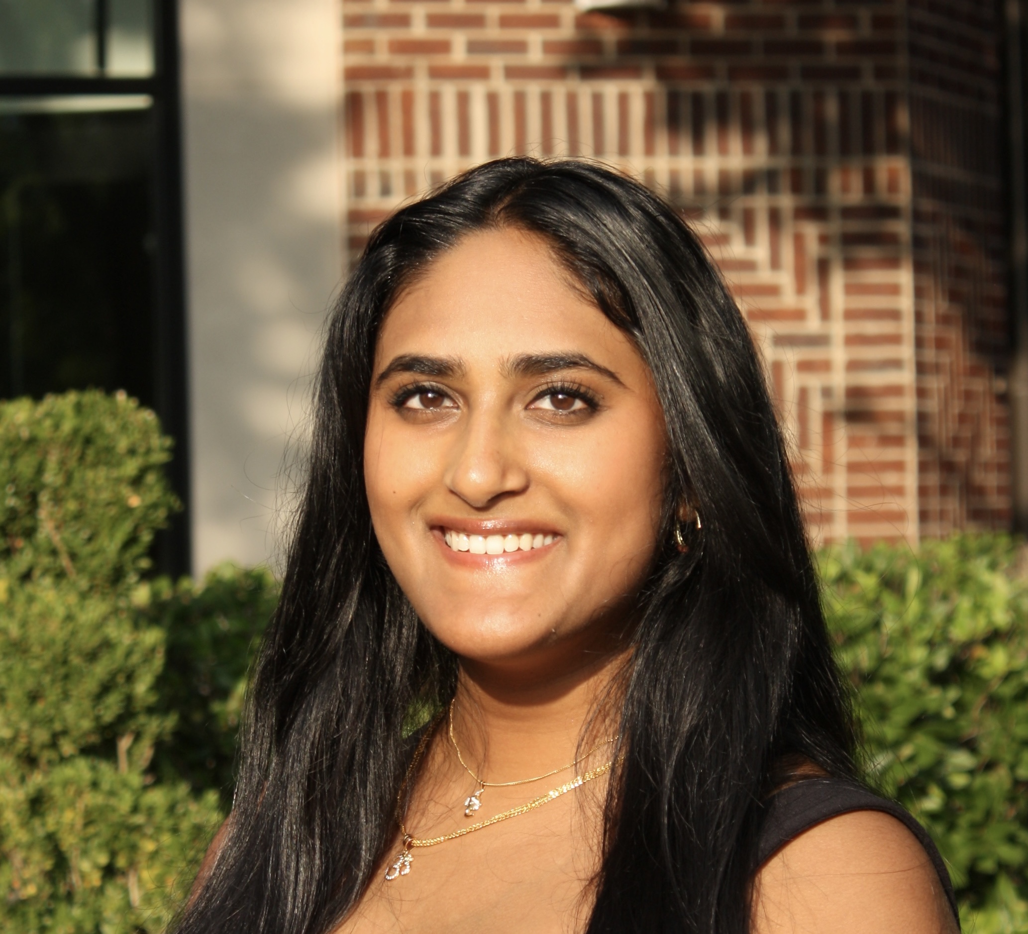 A young woman with center-parted dark hair stands against a background of bricks and green bushes. She is smiling and wearing a black tank top.