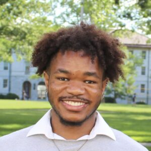 A Black Man With Twister Hair Above His Ears And A Light Beard Smiles At The Camera Against A Backdrop Of Green Trees And Sunny Lawn. He Is Wearing A Grey Sweater, White Collared Shirt And Gold Chain.