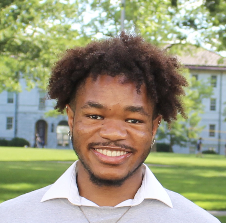 A Black man with twister hair above his ears and a light beard smiles at the camera against a backdrop of green trees and sunny lawn. He is wearing a grey sweater, white collared shirt and gold chain.