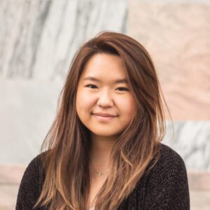 A Young Woman With Long Highlighted Side-parted Hair Stands Against A Grey And Pink Stone Wall. She Is Wearing A Black Cardigan.