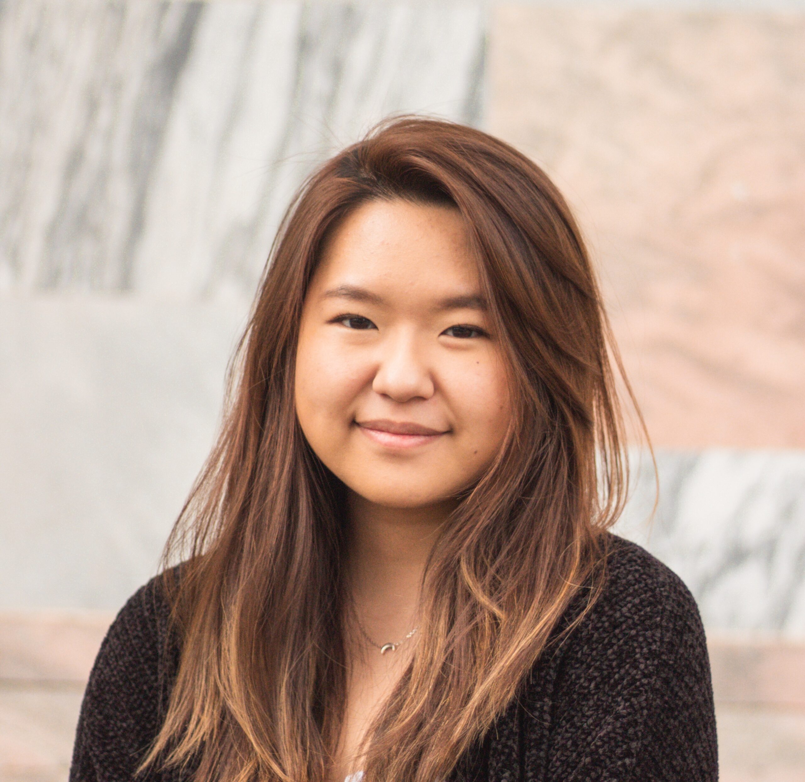 A young woman with long highlighted side-parted hair stands against a grey and pink stone wall. She is wearing a black cardigan.