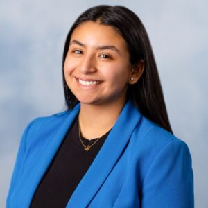 A Young Woman With Long Blak Hair Parted In The Middle Smiles At The Camera In A Sideways Pose. She Is Wearing A Black Round-necked Top And Royal Blue Blazer.