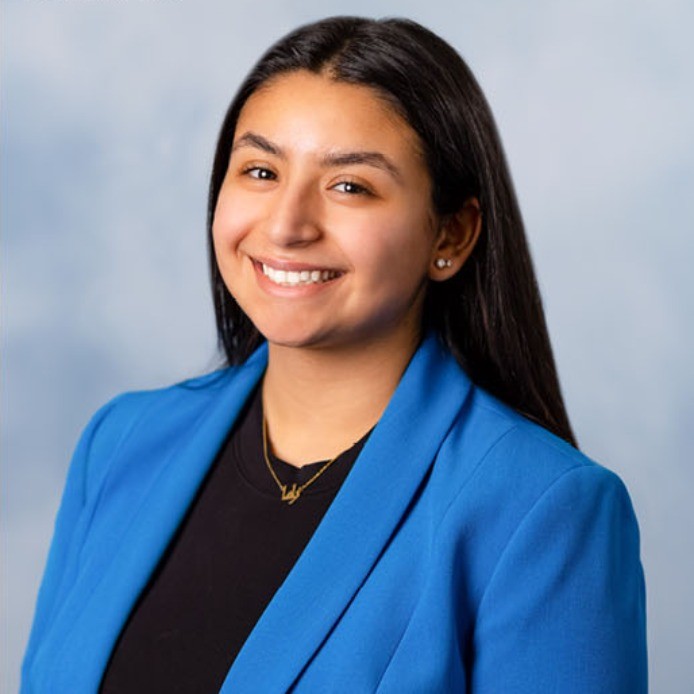 A young woman with long blak hair parted in the middle smiles at the camera in a sideways pose. She is wearing a black round-necked top and royal blue blazer.