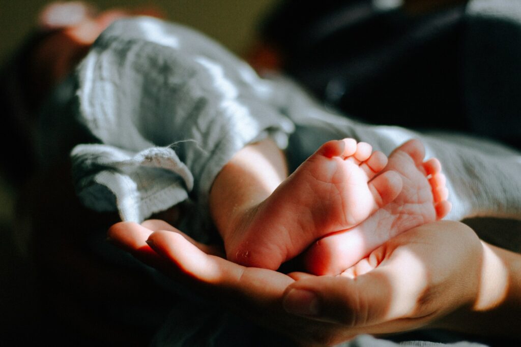 Against a black backdrop, a close-up shot shows the feet of a Caucasian baby wrapped in a light-blue blanket, being cradled by a Caucasian hand.