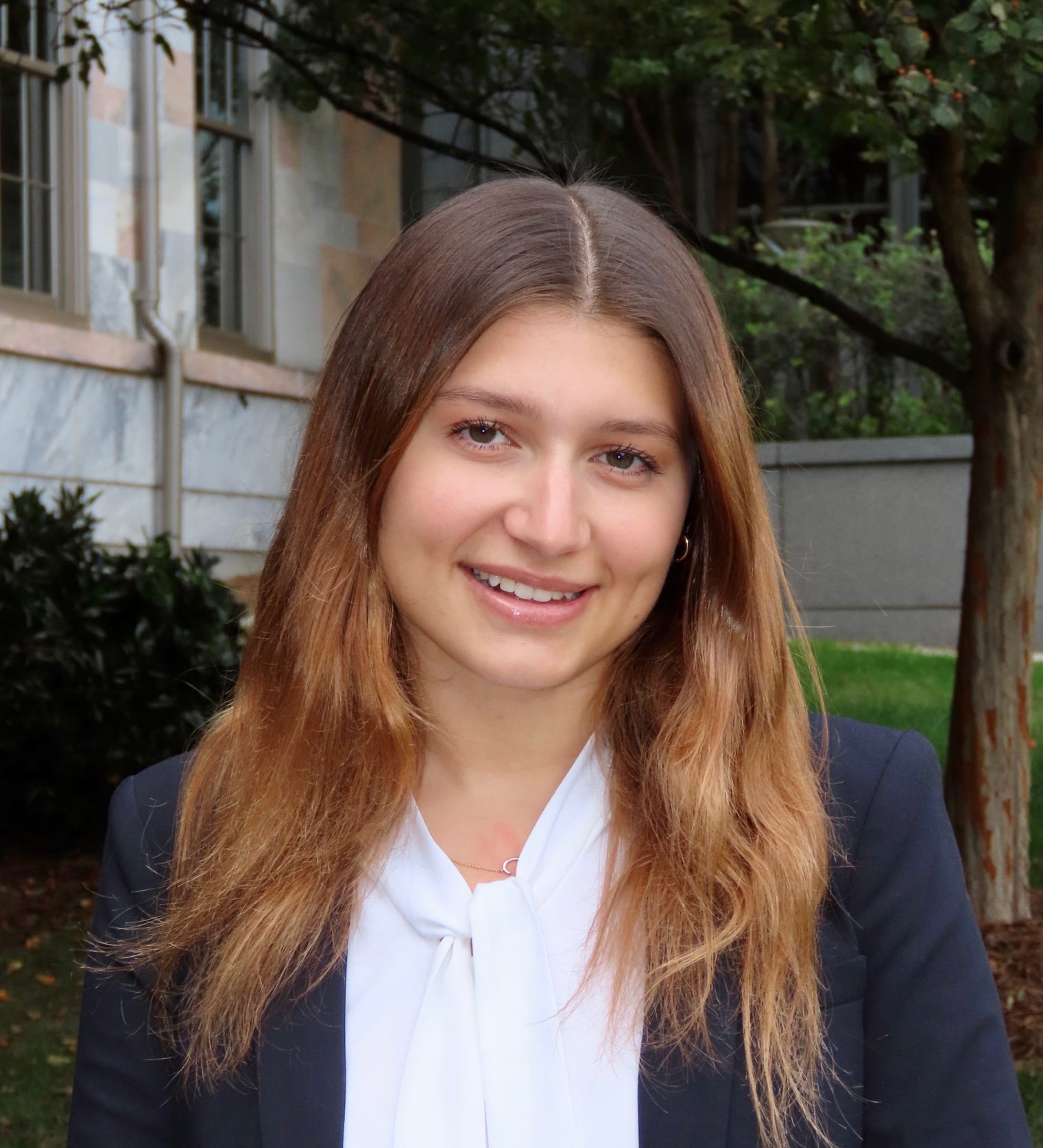 A young Caucasian woman with center-parted dark blonde hair smiles at the camera. She is wearing a blue blazer and a tie-front white blouse. She stands in front of a grey stone building with bushes and trees immediately behind her.