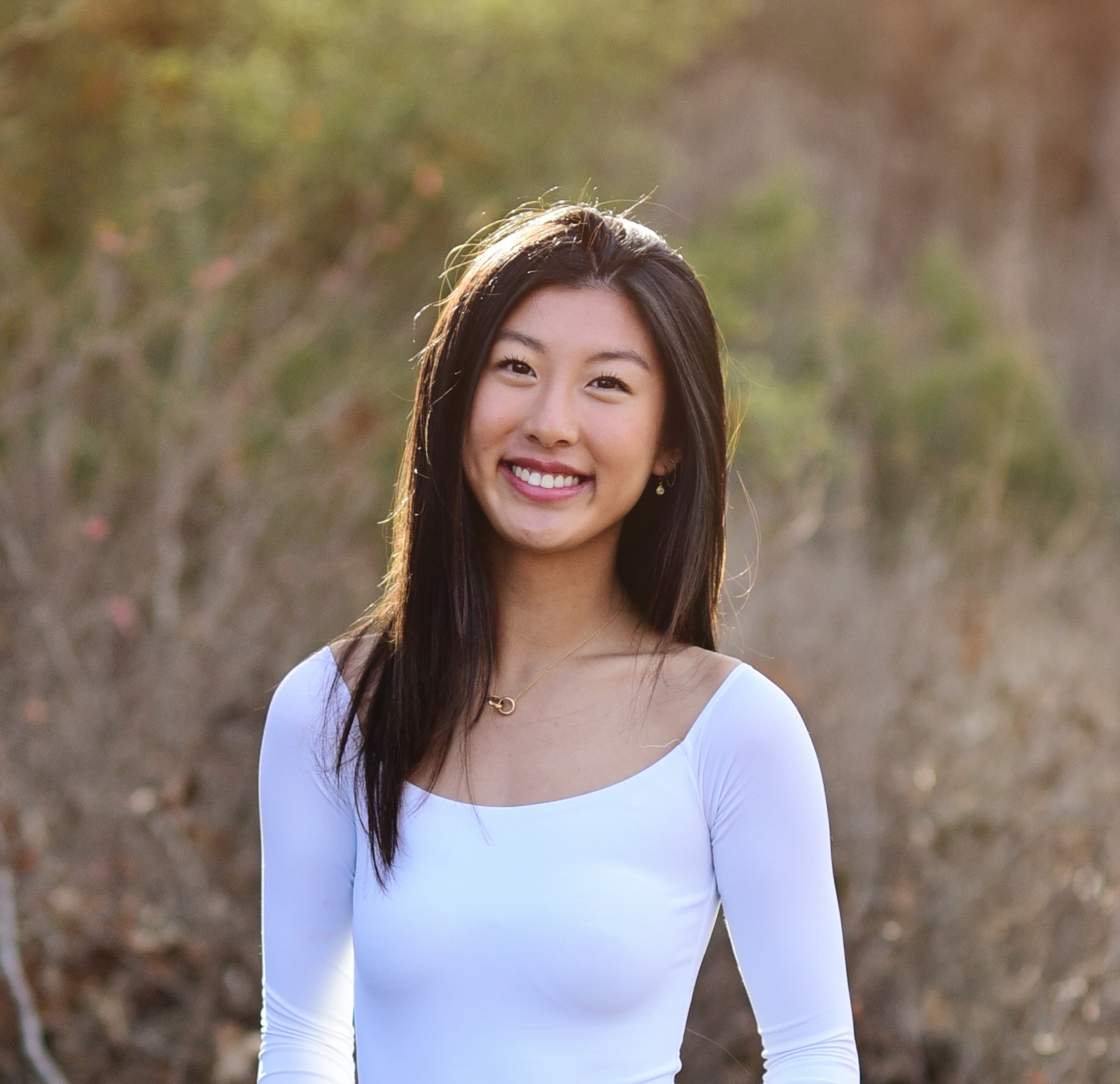 An Asian woman with a wide smile stands against an out-of-focus backdrop of vegetation. She is wearing a scoop-necked white top and a necklace of interlocking ring charms on a thin gold chain. The sun is coming in from the left side of the frame and highlighting her straight dark hair.