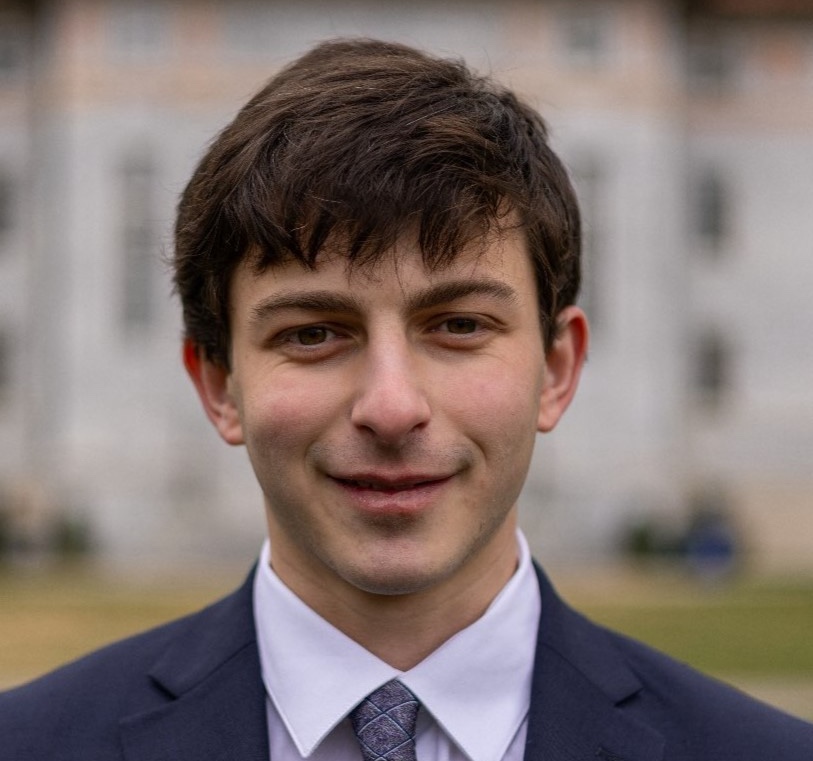 A dark-haired young man with a Caucasian skin tone smiles slightly at the camera. He has dark hair that lies across his forehead. He is wearing a navy blazer, whie shit and dark tie, and is posed against an out-of-focus building of grey and pink granite.
