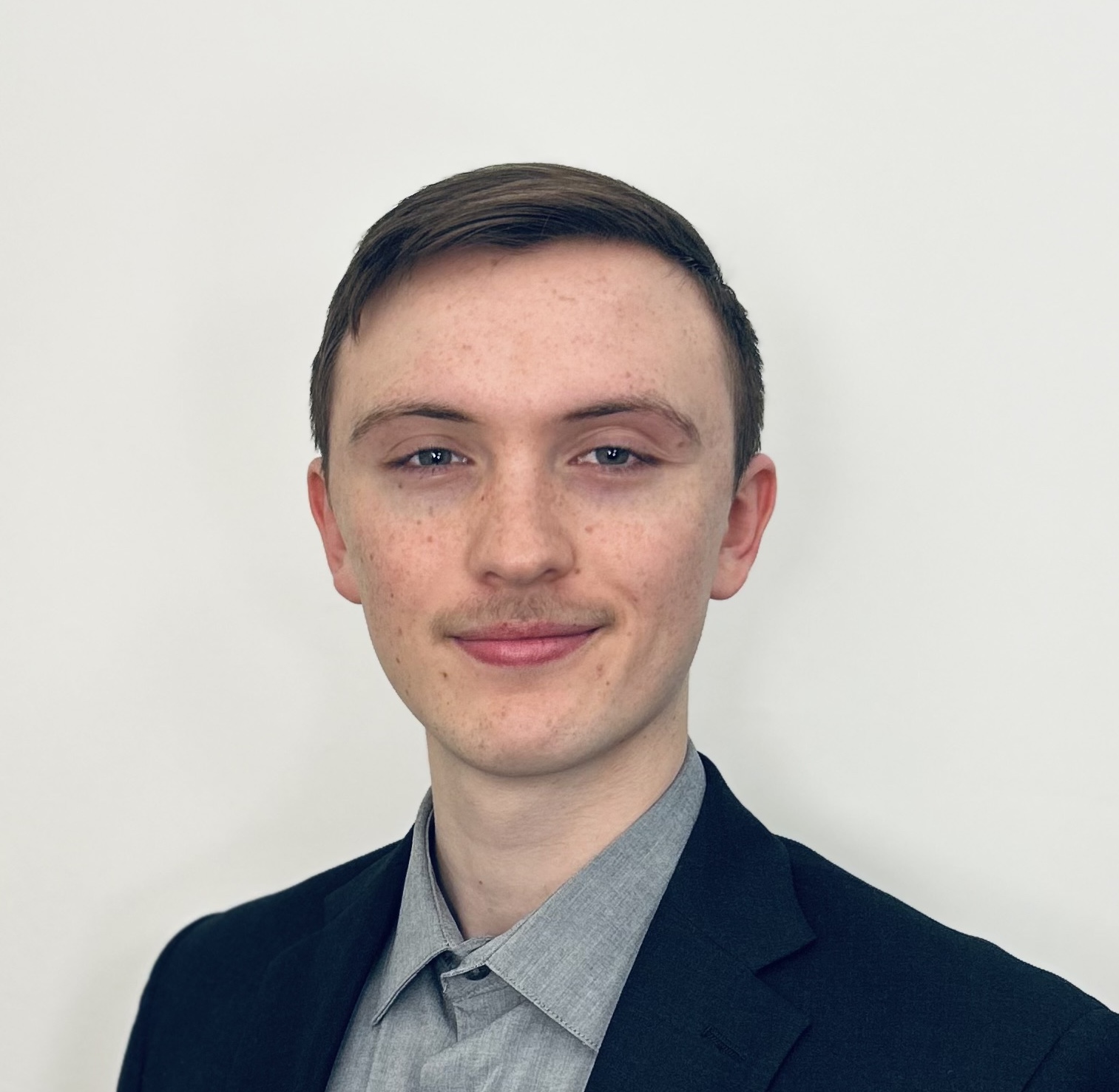 A young man smiles at the camera. He is pale-skinned and lightly freckled and is posed against an off-white background, wearing a grey short closed at the neck with no tie, and a navy blazer. His brown hair is parted and combed to one side, and he sports a trimmed moustache.