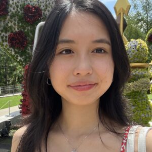 An Asian Woman With Center-parted Dark Hair That Falls Below Her Shoulders Looks Slightly To The Right Out Of Camera Range. She Is Wearing A Strappy Top And A Gem Pendant. She Is Standing In Sunlight Against A Background Of Garden Installations Made Of Flowers.