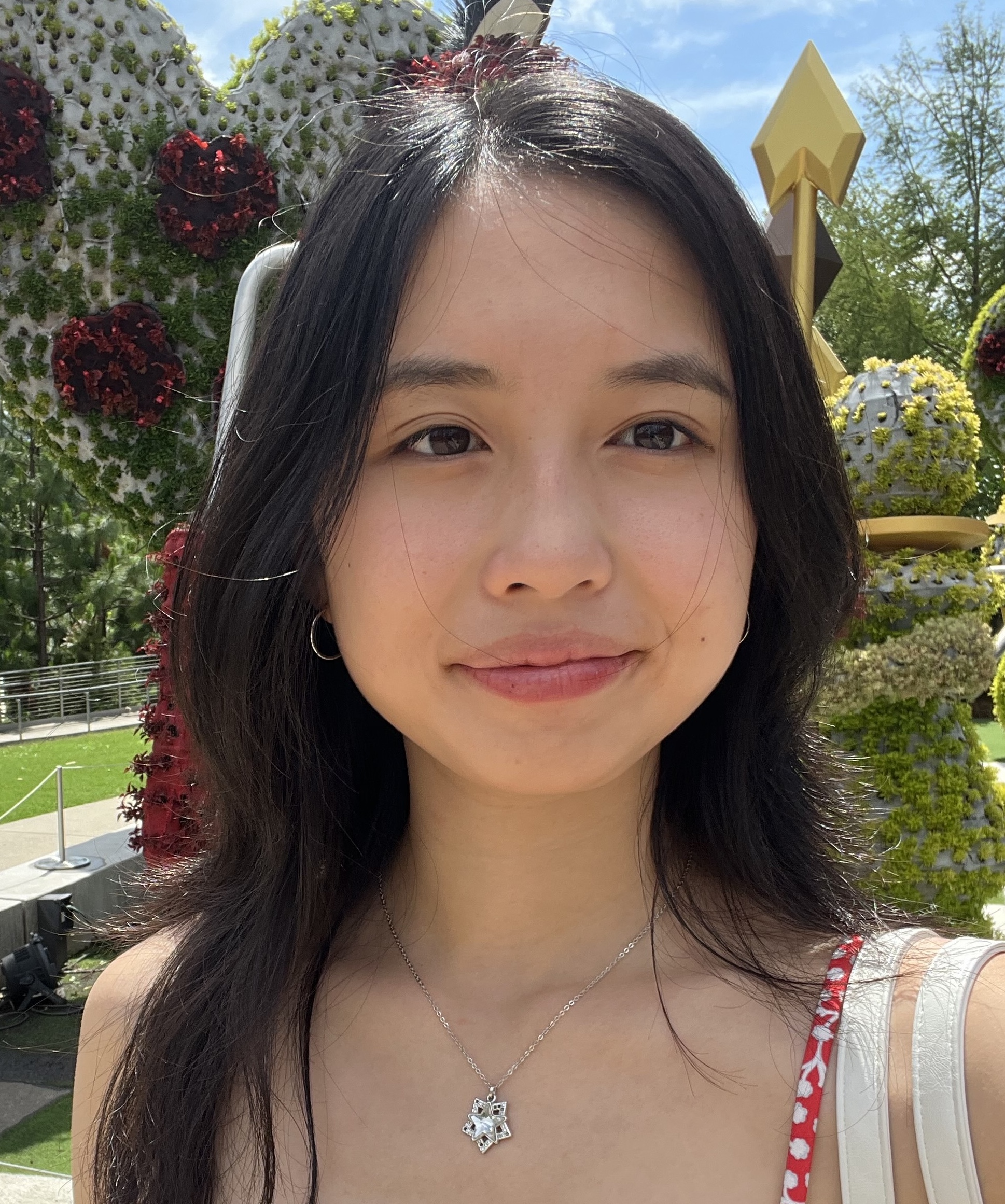 An Asian woman with center-parted dark hair that falls below her shoulders looks slightly to the right out of camera range. She is wearing a strappy top and a gem pendant. She is standing in sunlight against a background of garden installations made of flowers.