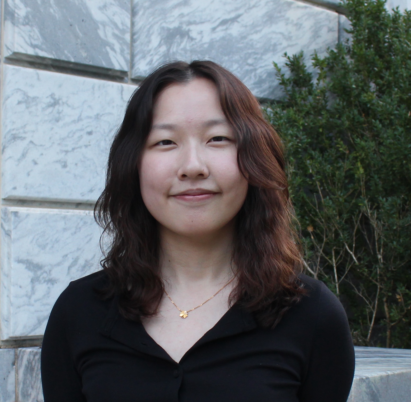 A young Asian woman with wavy center-parted hair that stops at her collarbones smiles with a closed mouth against a grey stone building. She is wearing a tiny bright-gold charm on a narrow chain and a v-necked black top. There is a green boxwood bush behind her left shoulder.