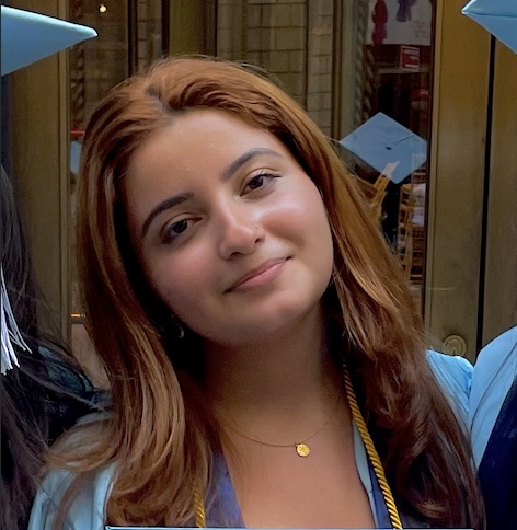 A young woman with center-parted long auburn hair tilts her head and smiles slightly at the camera. She appears to be attending a graduation ceremony because people wearing bright-blue mortarboards are leaning in to both sides of the frame and reflected in a surface behind her head. A gold chain with a tiny medallion rests in her suprasternal notch.