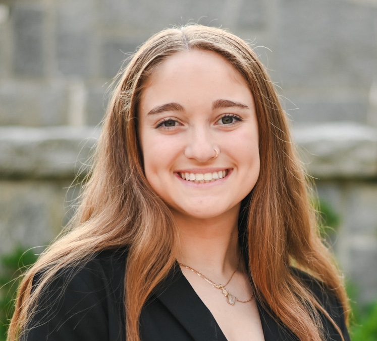 A young white woman with straight eyebrows and center-parted light red hair smiles at the camera. Her long hair cascades past her shoulders over a black blazer. She wears a gold nostril ring and two narrow gold chain necklaces and is posed against a gray stone wall that is out of focus.