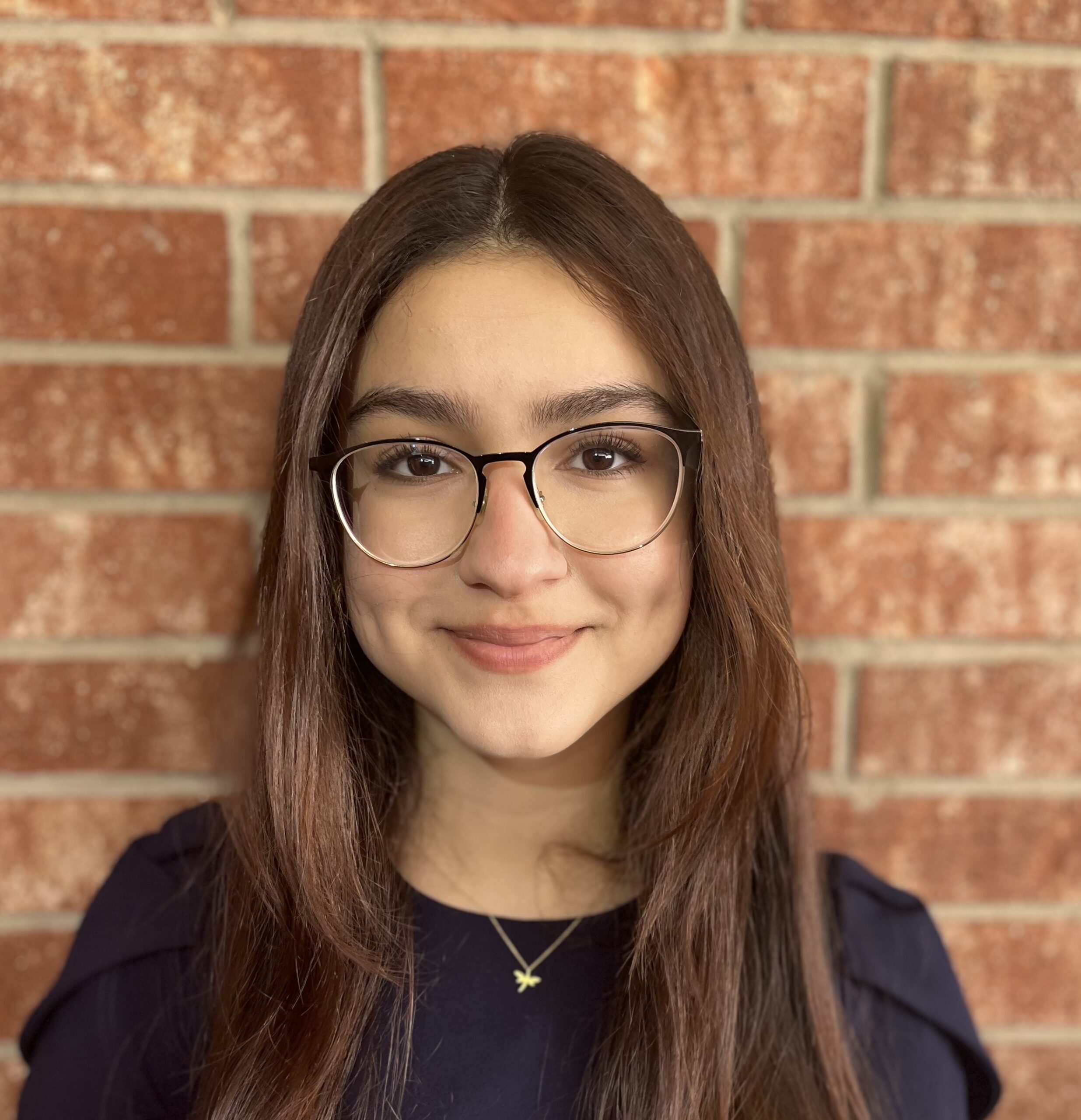 A young woman with medium-toned skin and center-parted brown hair smiles at the camera against a backdrop of reddish bricks. She is wearing a dark-blue top with shoulder pleating, a charm on a thin gold neck chain, and glasses with rounded frames that are dark along the top edge.
