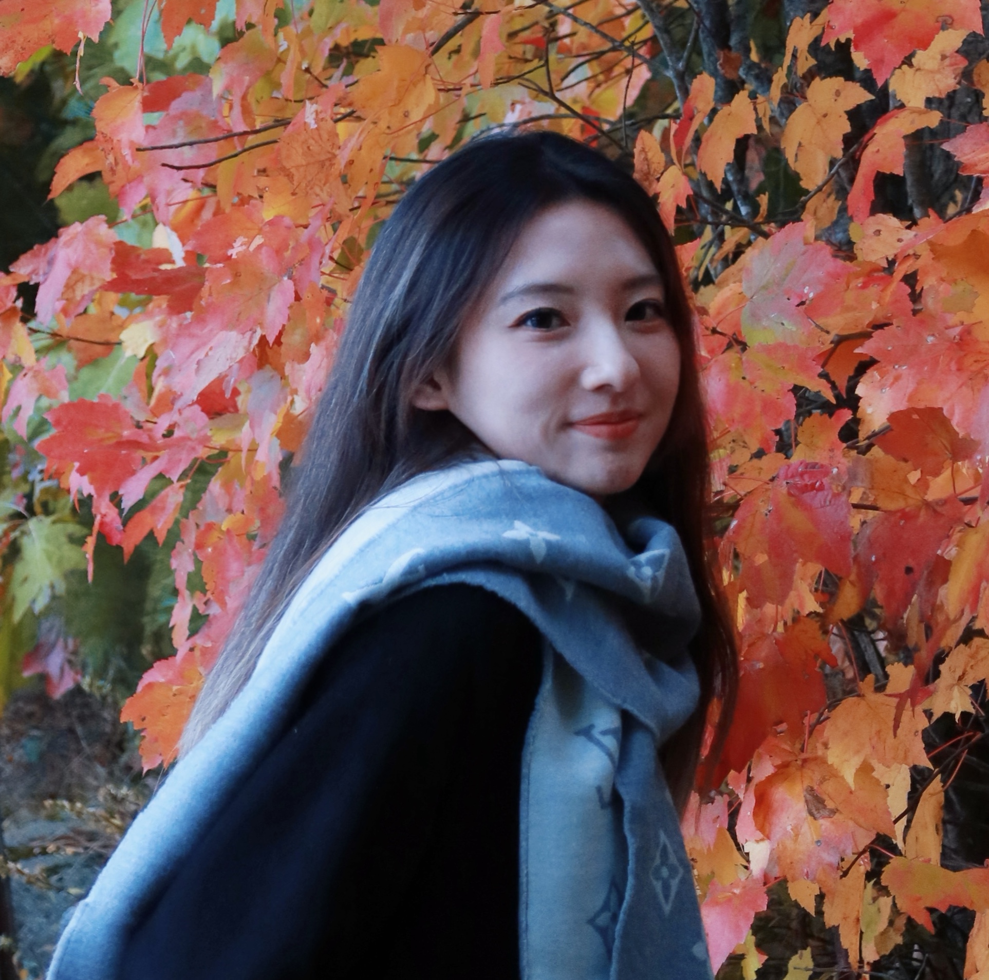 A young Asian woman with long dark hair looks over her shoulder at the camera against a backdrop of autumn leaves. She is wearing a dark coat and a pale blue Louis Vuitton scarf. She is smiling slightly.