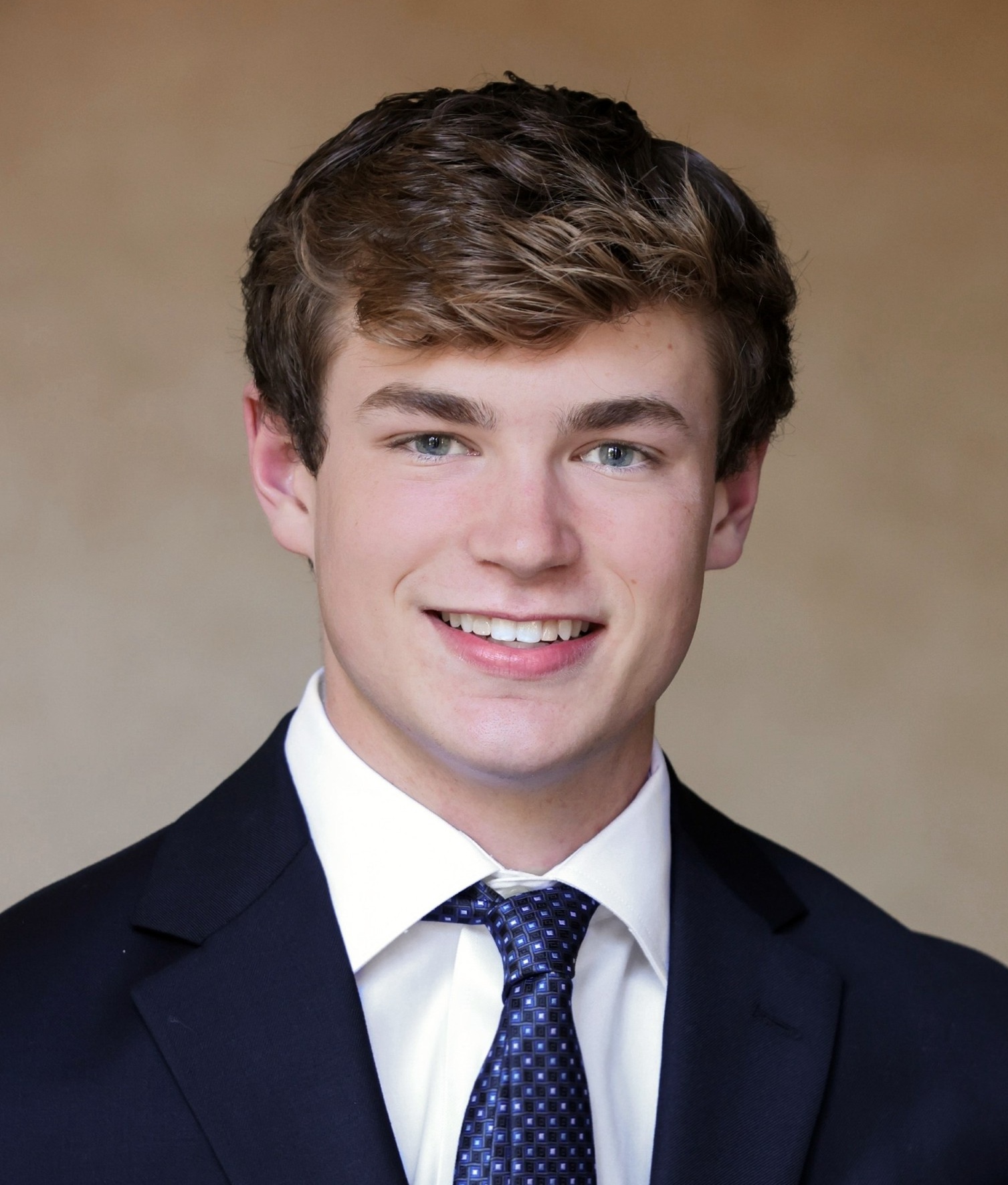 A young white man with thick dark-blond hair poses against a mottled ochre background. He is smiling and wearing a white shirt, navy blazer, and patterned dark blue tie knotted in a four-in-hand knot.