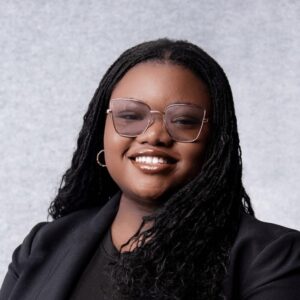 A Dark-skinned Young Woman With Long Dark Hair Smiles Against A Grey Background. She Is Wearing A Black Jacket, A Black Top, And Large Tinted Glasses.