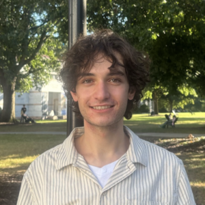 A Young Fair-skinned Man With Curly Hair Reaching The Tops Of His Ears Smiles Against A Landscape Of Leafy Trees And Slanting Sunlight. He Is Wearing A White T Under An Open-collared Shirt Striped In Ochre And White.