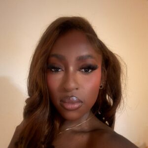 A Dark-skinned Young Woman With Cascading Chestnut Hair Stands Against A Beige Backlit Background. She Is Wearing A Narrow Gold Necklace.